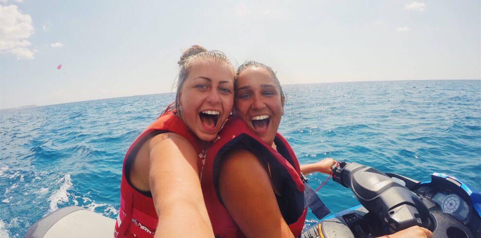 Tourists enjoying watersports in Bali, including jet skiing, parasailing, and banana boat rides against a backdrop of clear blue water and a sunny sky